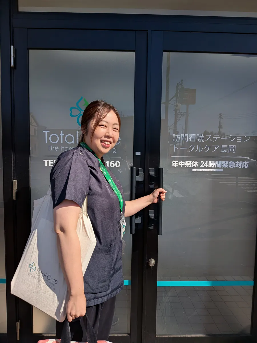 訪問バッグを肩に、ステーションの入口で振り返って笑う看護師