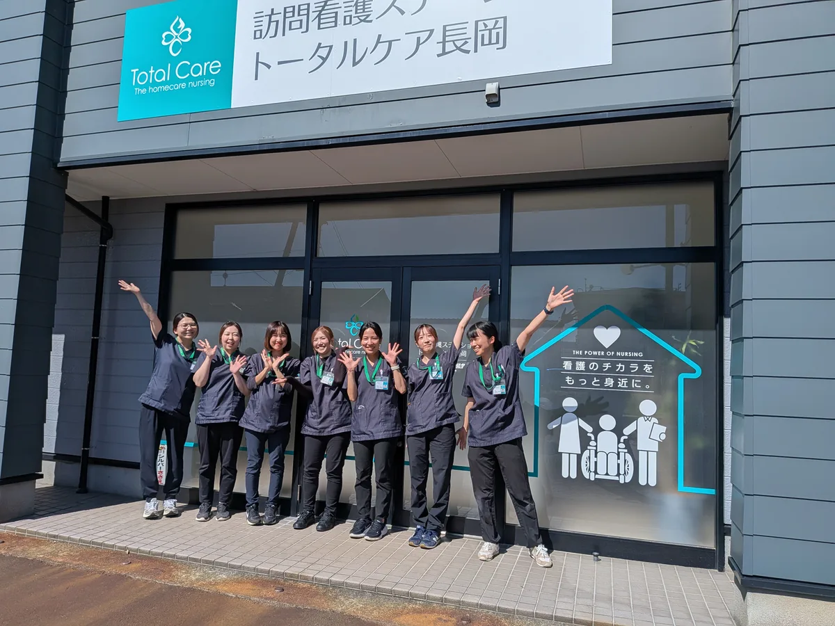 トータルケア長岡の看板の前で、7人の看護師が手を振って笑う
